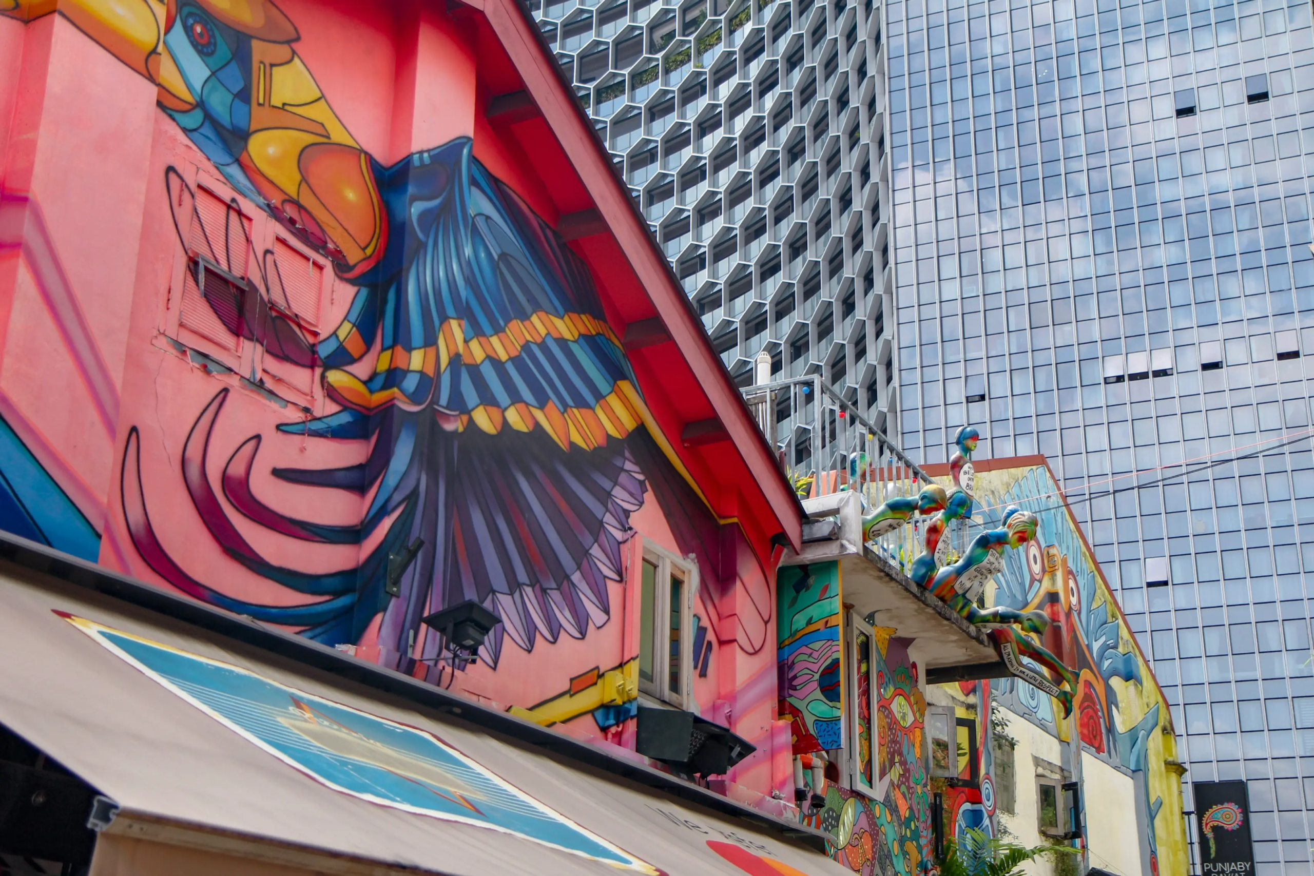 A vibrant, colorful mural of a bird adorns the side of a pink building, juxtaposed against a modern skyscraper with a glass and honeycomb facade. The scene captures a striking contrast between the expressive street art of a traditional neighborhood and the sleek architecture of a contemporary city.