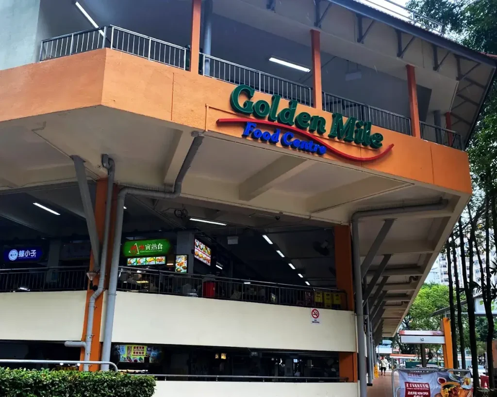 Golden Mile Food Centre exterior, featuring bright signage. The building has two levels and vibrant colors. Trees nearby, creating a lively atmosphere.