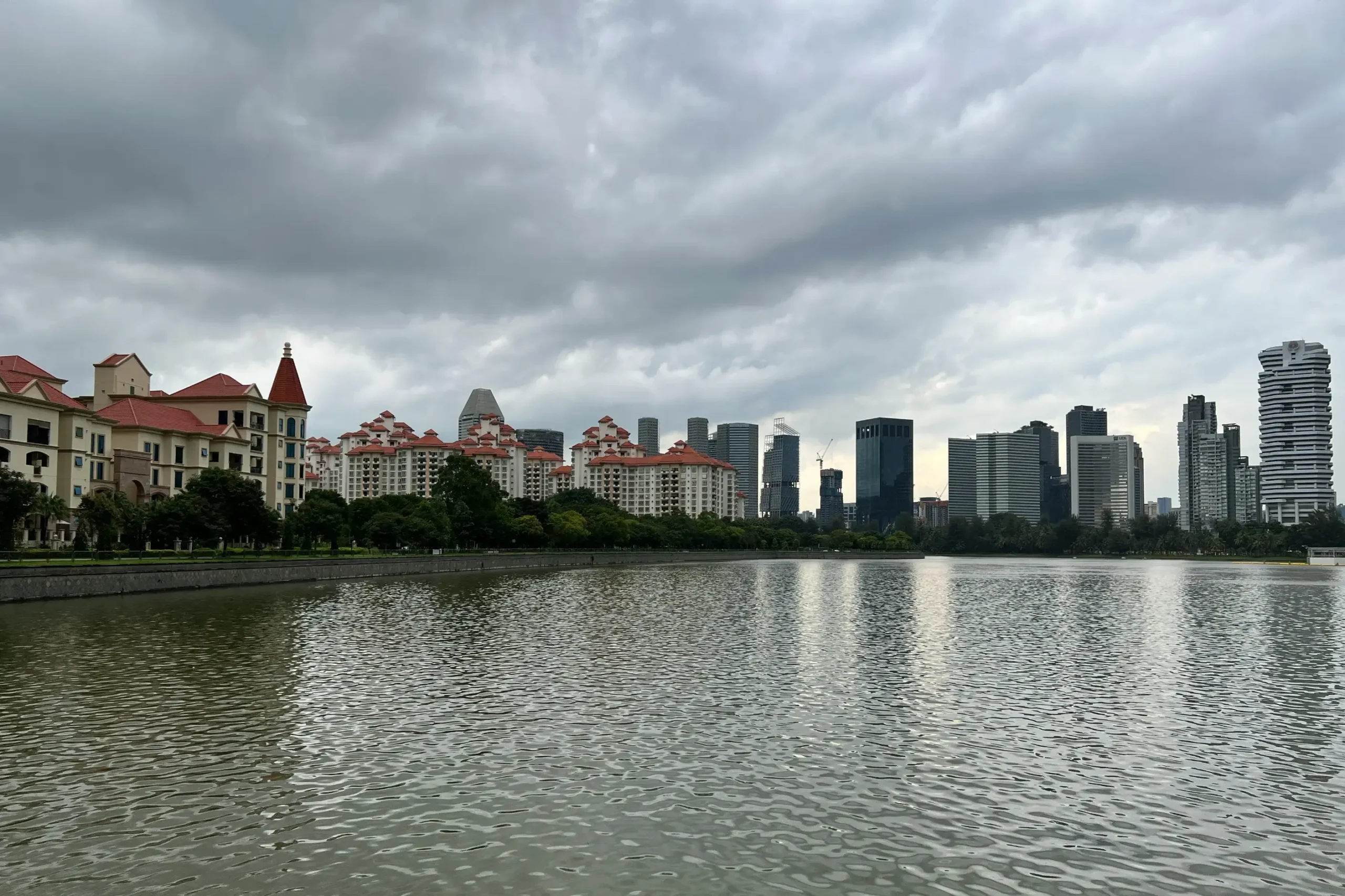 This image captures a wide view of a city skyline across a calm body of water under a heavy, overcast sky. Modern high-rise buildings and red-roofed apartment complexes line the far bank, framed by a row of lush green trees.