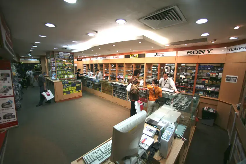 This image shows the interior of a well-lit camera shop, featuring glass display cases filled with photography equipment and various brand signs along the walls. Several customers are interacting with staff behind the counters, while old-fashioned monitors and keyboards are visible in the foreground.