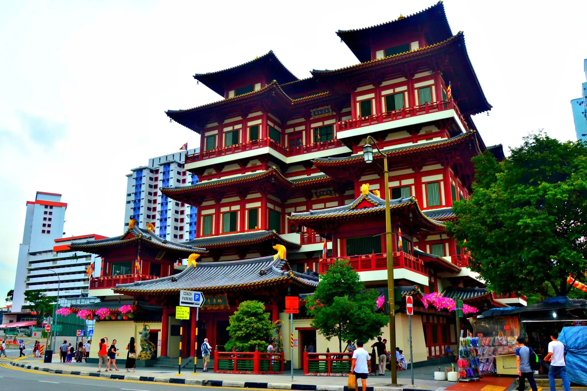 The Buddha Tooth Relic Temple stands as a magnificent multi-story landmark in Singapore, featuring vibrant red walls and traditional Tang-style architectural details. Pedestrians walk along the adjacent sidewalk near street signs and colorful floral decorations that line the base of the ornate structure.