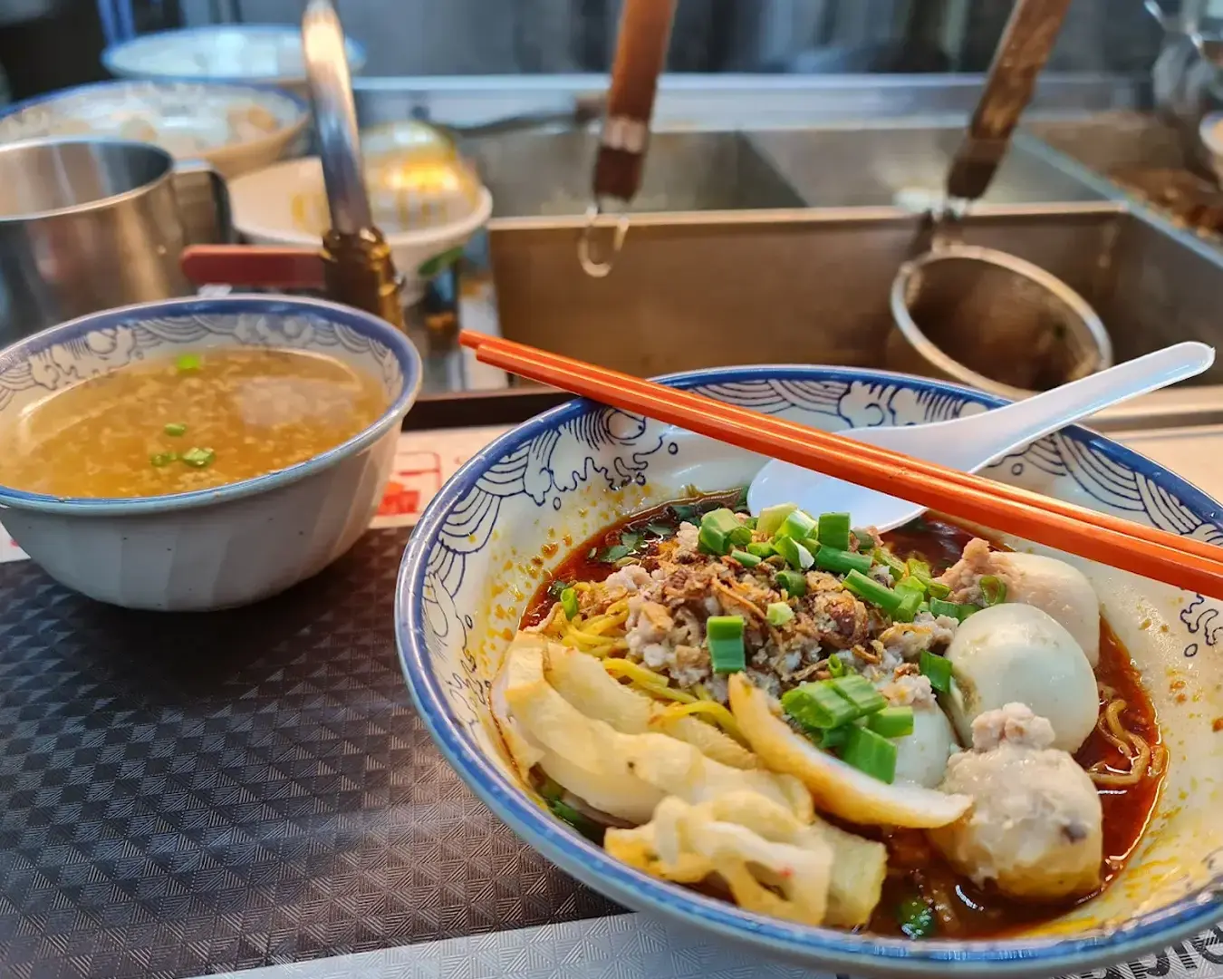 A vibrant bowl of spicy noodle soup garnished with green onions and chopsticks sits beside a bowl of clear broth with herbs, set on a serving tray.