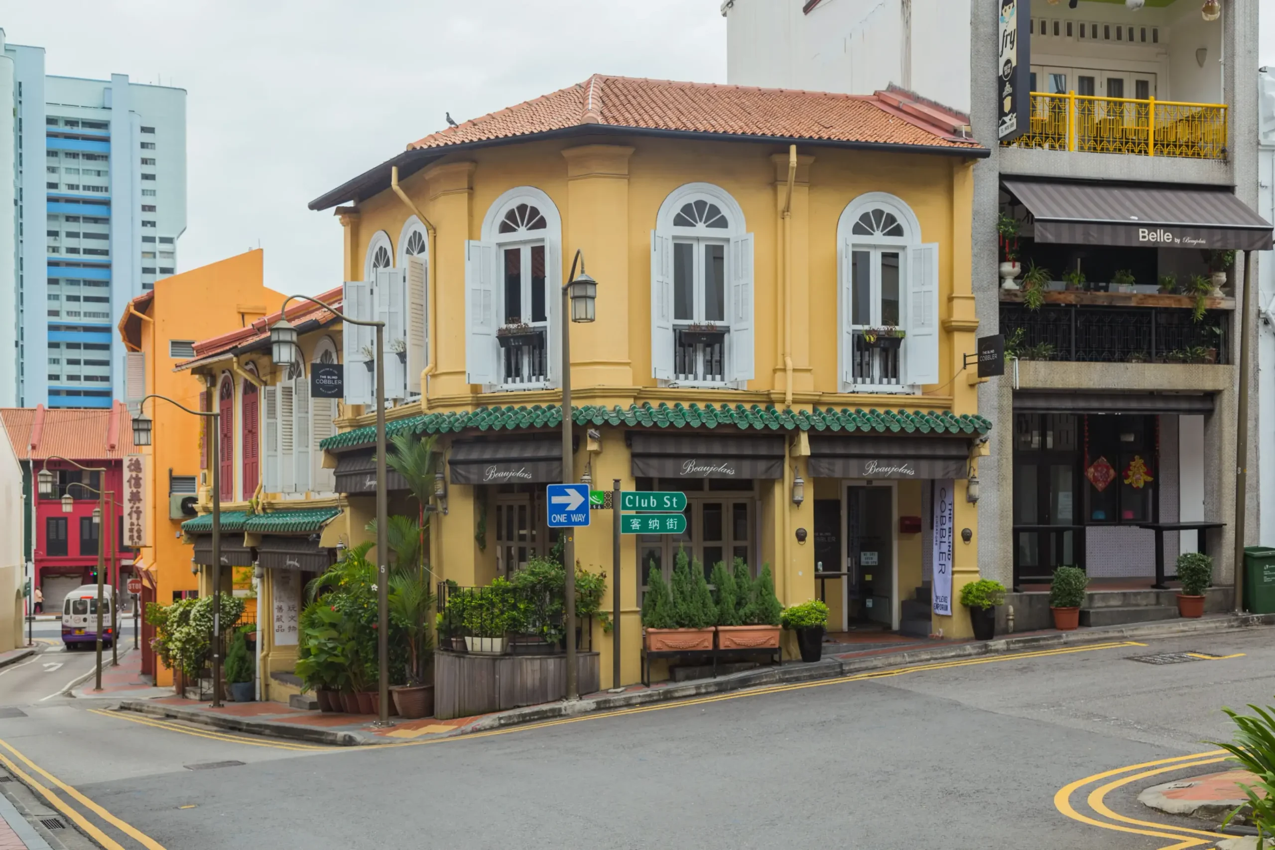 This image captures a vibrant street corner in Singapore's Chinatown, featuring a distinctive yellow heritage shophouse at the intersection of Ann Siang Hill and Club Street. The scene showcases a blend of traditional architecture with modern urban elements, characterized by arched windows, decorative green tiling, and various commercial storefronts.