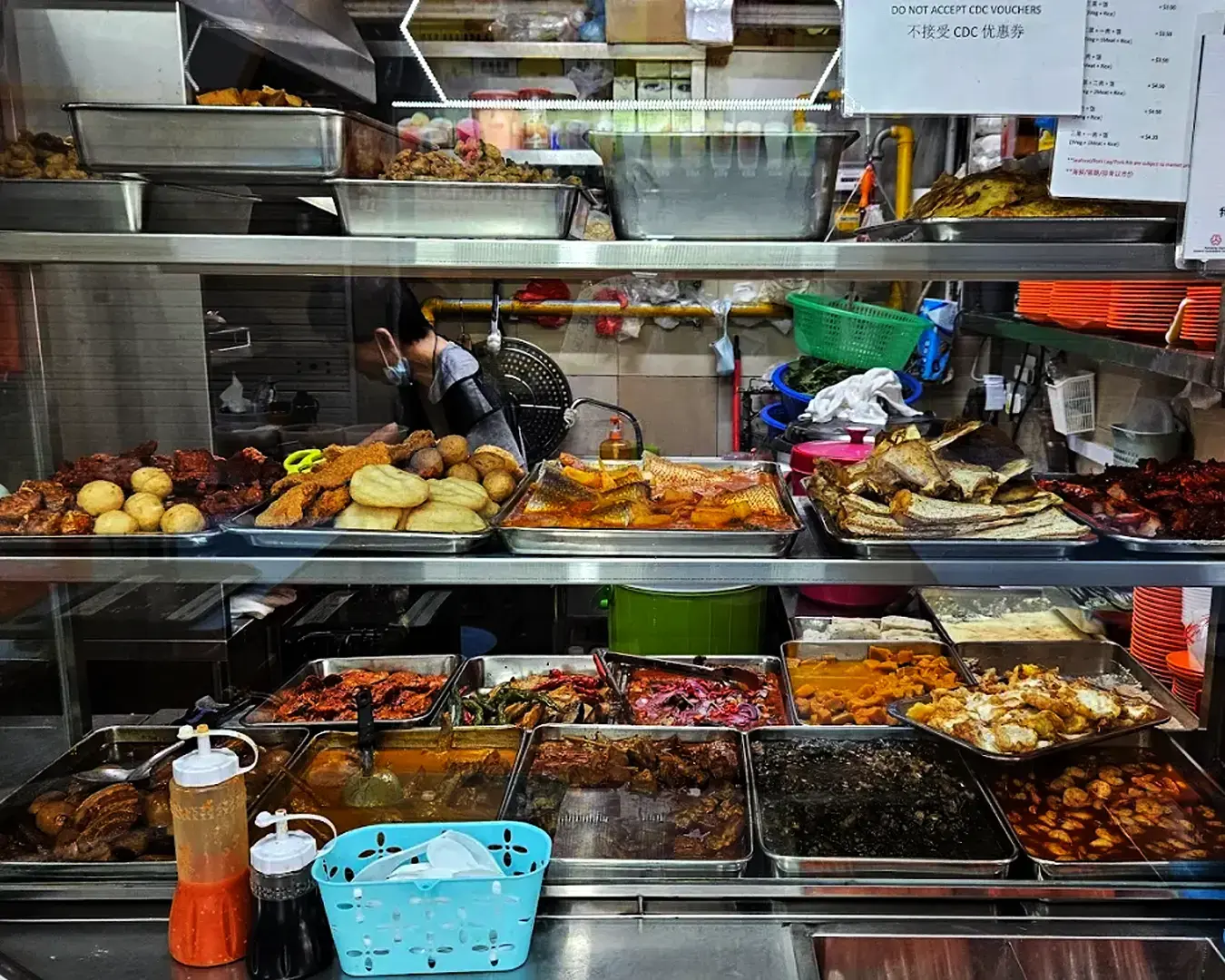 A vibrant food stall displays an array of colorful dishes in metal trays. Visible are meats, fish, and vegetables under warm lighting, conveying a bustling, appetizing scene.