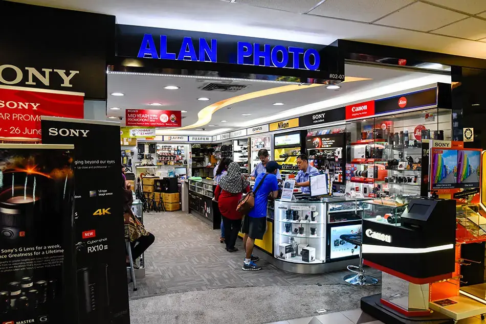 The image shows the brightly lit storefront of Alan Photo, a camera shop featuring prominent signage for brands like Sony, Nikon, and Canon. Inside, staff members in blue shirts assist customers at a glass display counter filled with photography equipment and accessories.