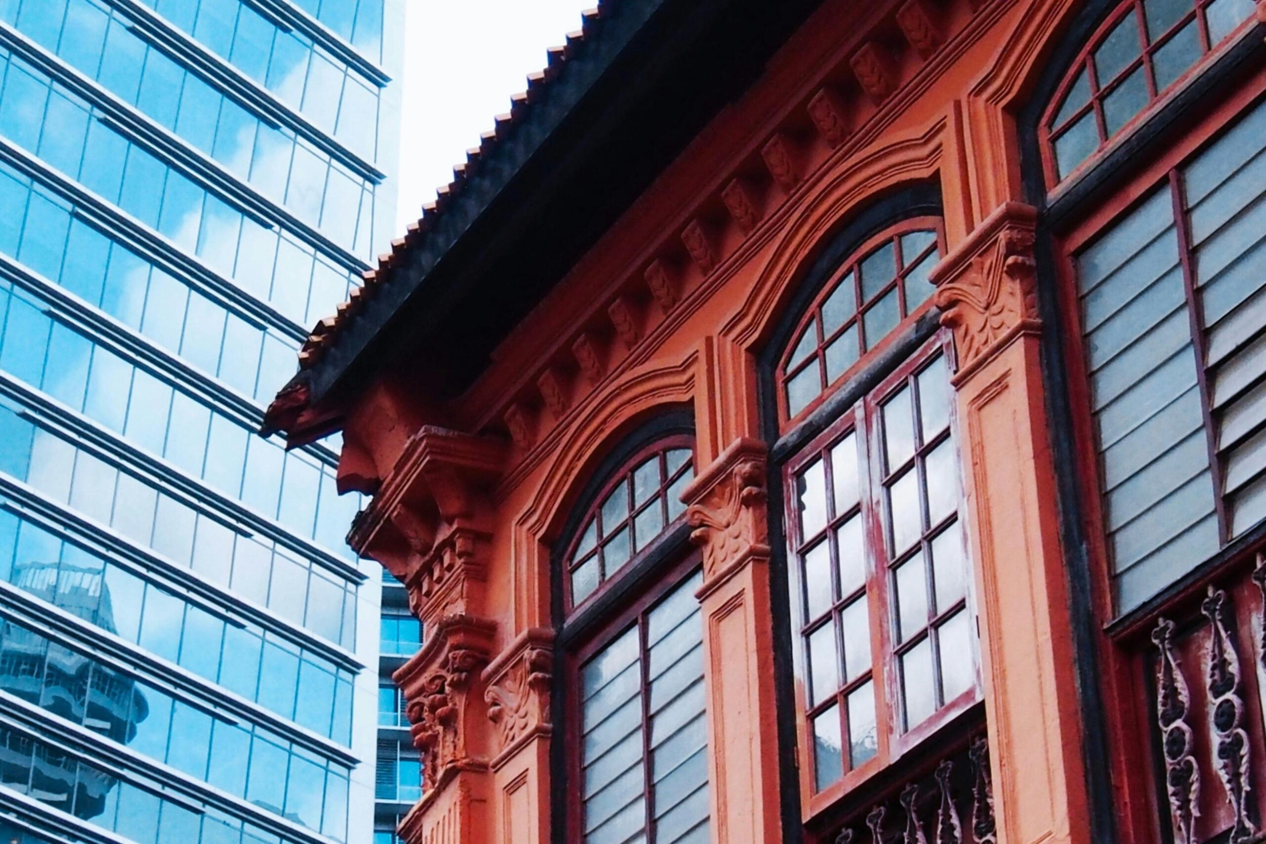 This image captures a vibrant, orange shophouse featuring ornate architectural details and traditional arched windows. It stands in stark contrast to the sleek, blue glass facade of a modern skyscraper rising in the background.
