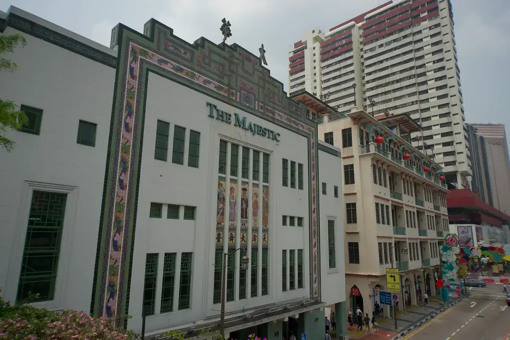 The Majestic building stands as a prominent historical landmark in Singapore, featuring an ornate facade with colorful mosaic tiles and Chinese opera figures. Set against a backdrop of modern high-rises and bustling city streets, the architecture highlights the blend of traditional heritage and contemporary urban life.