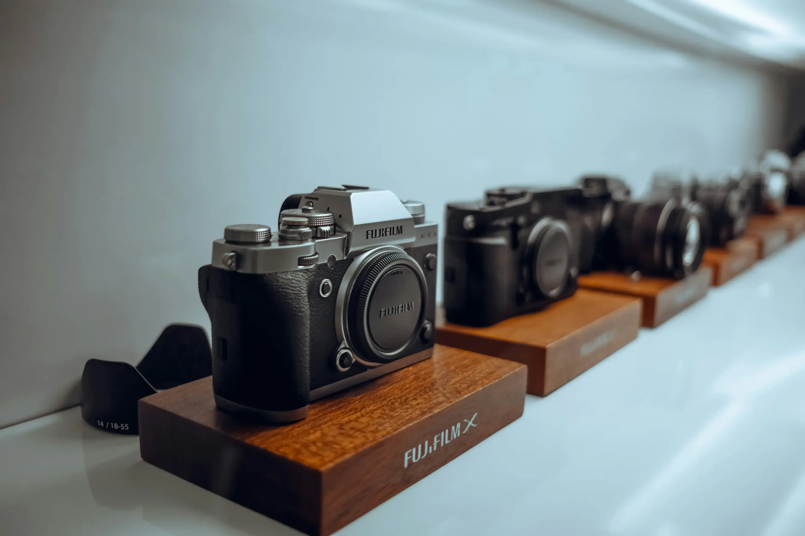 This image features a row of Fujifilm mirrorless cameras displayed on individual wooden blocks along a sleek, white shelf. The focus is sharp on a silver and black model in the foreground, while the others recede into a soft, shallow depth of field.