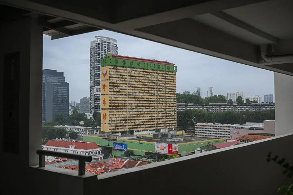 This shot frames the iconic, brightly colored People's Park Complex in Singapore through the geometric opening of a concrete structure. The yellow and green facade of the building stands out against a cloudy sky, surrounded by a mix of modern skyscrapers and traditional red-roofed shophouses.