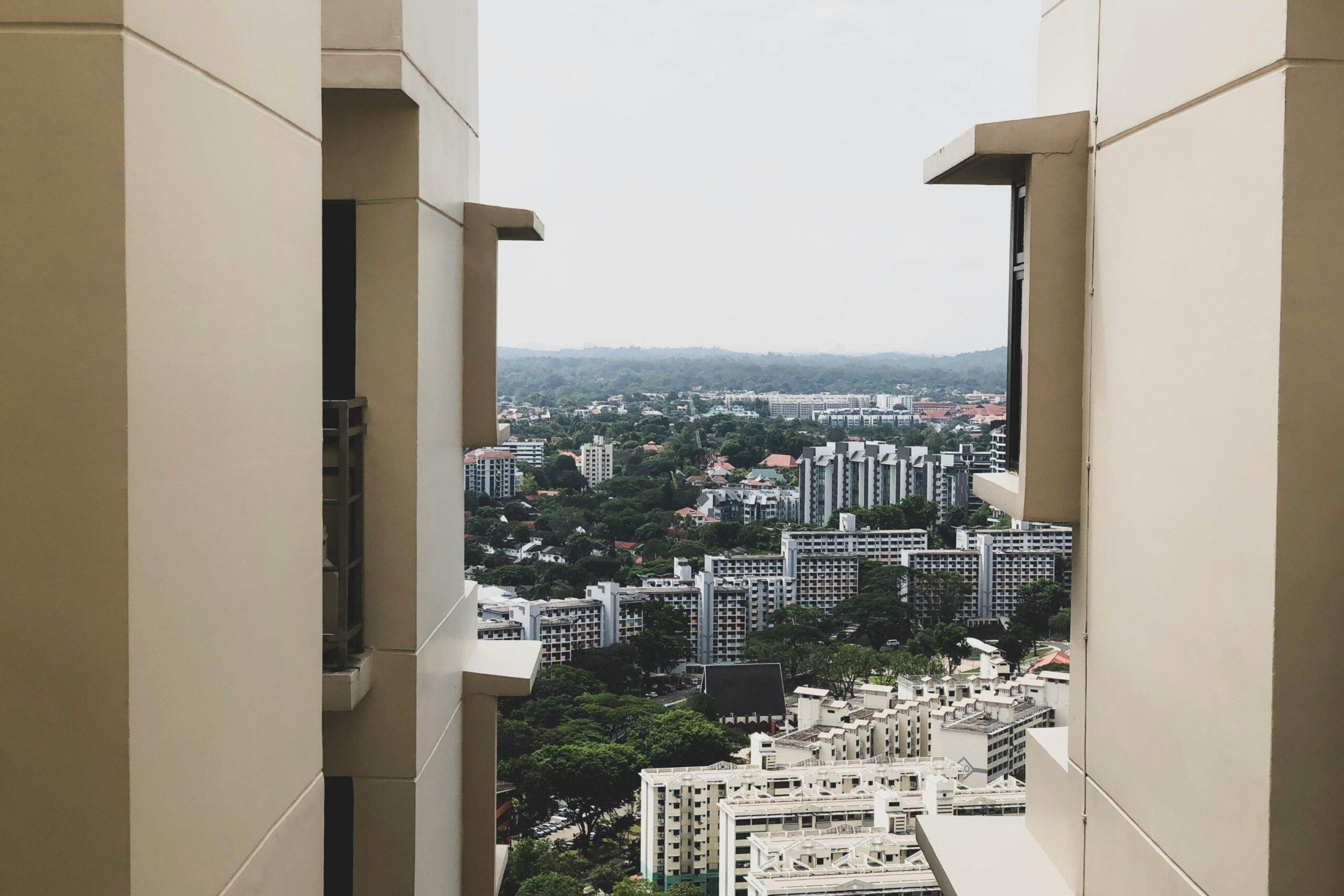 This high-angle shot frames a dense urban landscape between the neutral-toned walls of two high-rise buildings. Beyond the immediate structures, a sprawling residential area transitions into a lush, green horizon under an overcast sky.