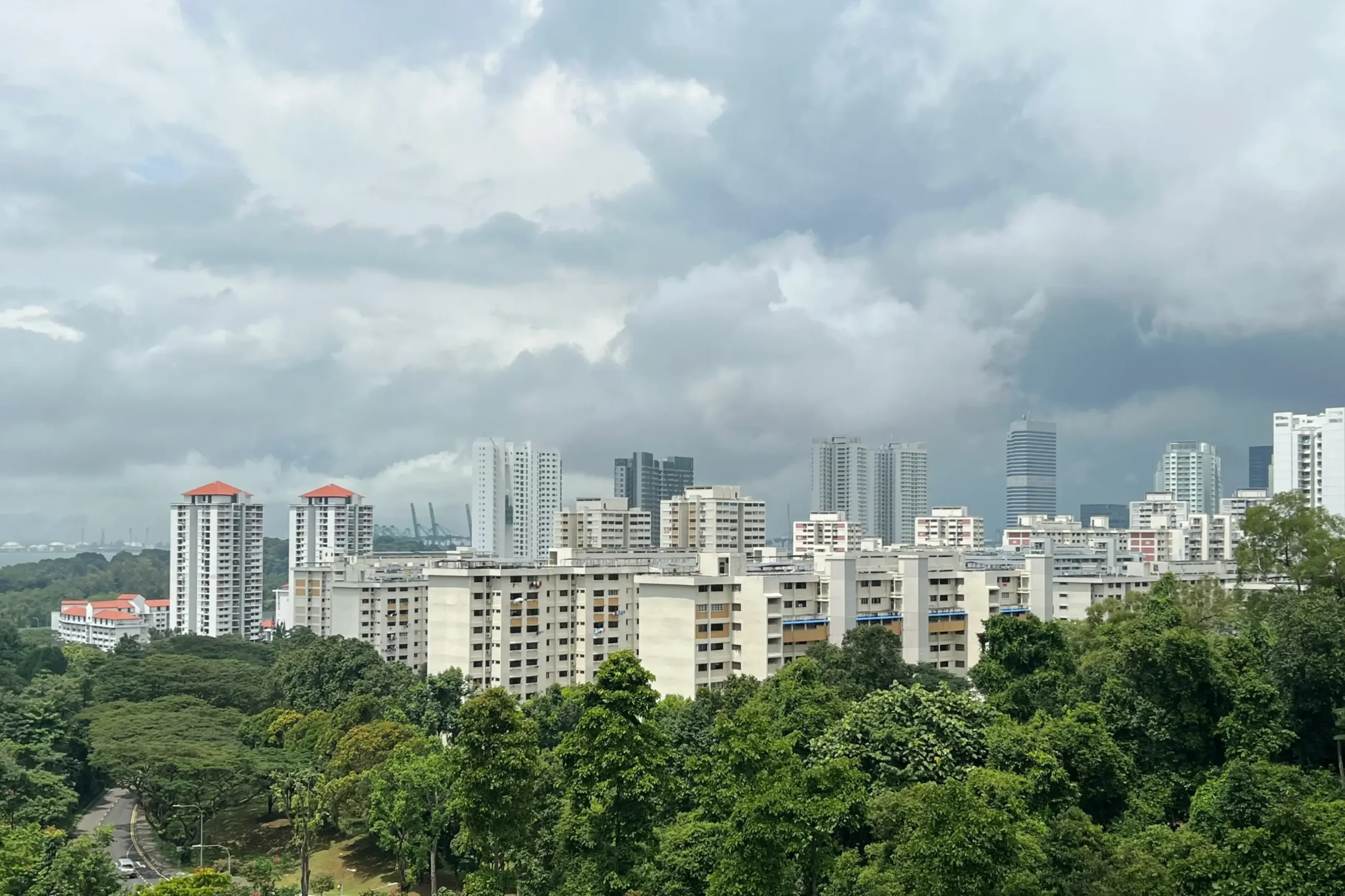 This wide shot captures an expansive urban landscape in Singapore, featuring a dense cluster of white high-rise residential buildings nestled behind a lush green forest. Above the cityscape, a vast, overcast sky filled with heavy grey clouds suggests an approaching tropical rainstorm.