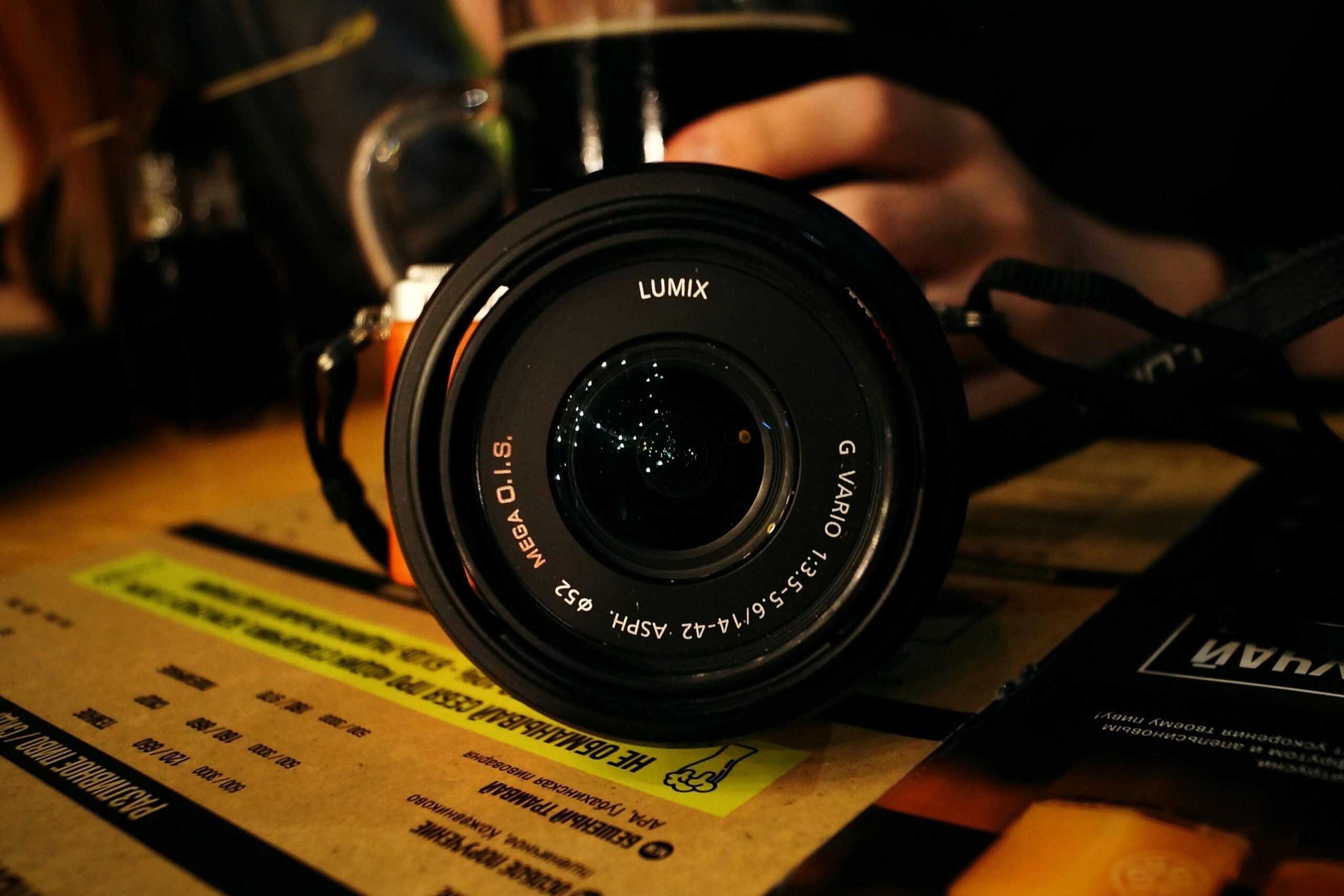 A close-up shot captures a black Lumix camera lens resting on a table with text-covered documents. The lens reflects subtle ambient lights, while the background remains softly blurred and out of focus.