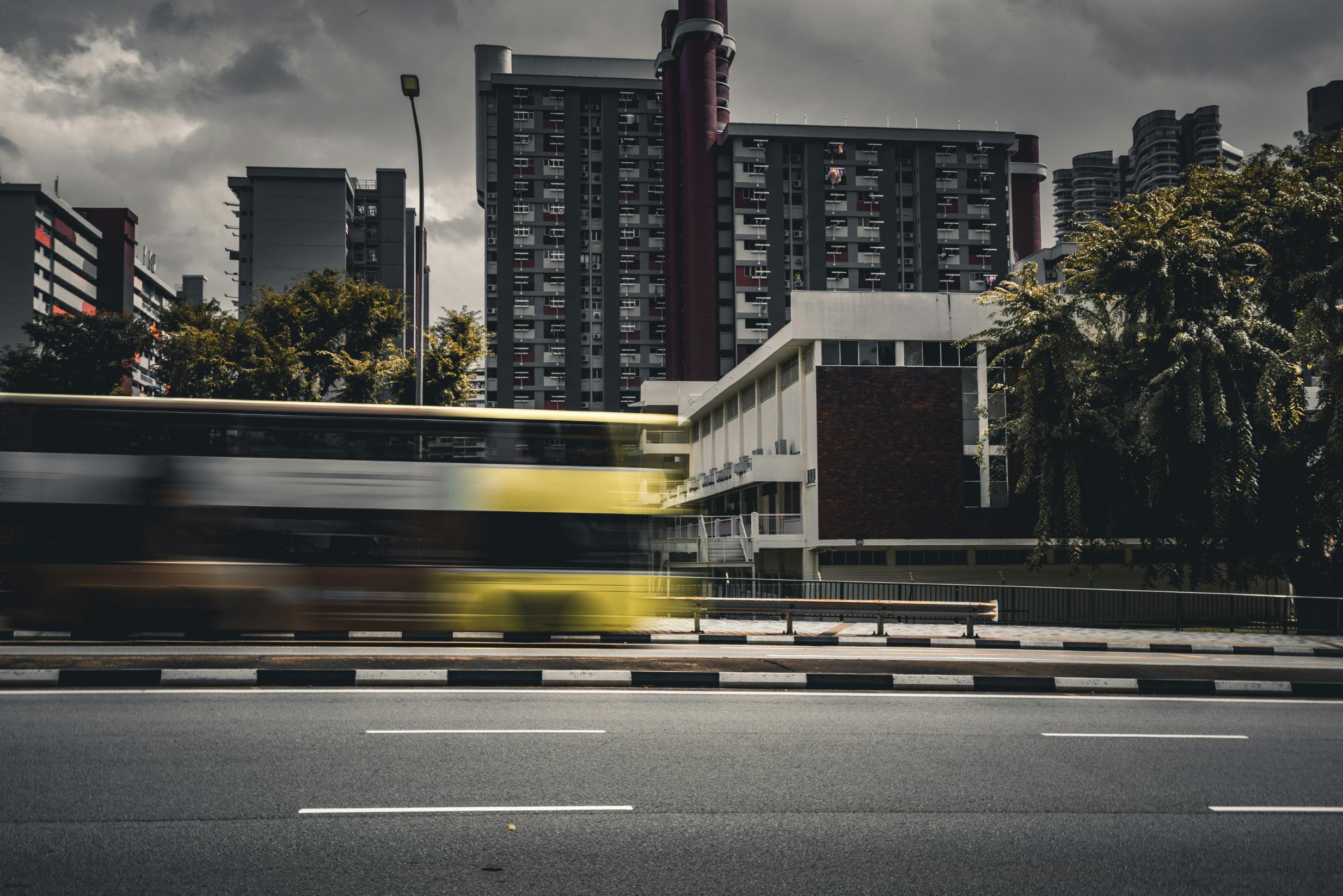 A yellow and white double-decker bus streaks across the foreground of a dark, urban landscape, captured with a motion blur that emphasizes speed. In the background, looming residential high-rises stand under a heavy, overcast sky, creating a moody and industrial atmosphere.