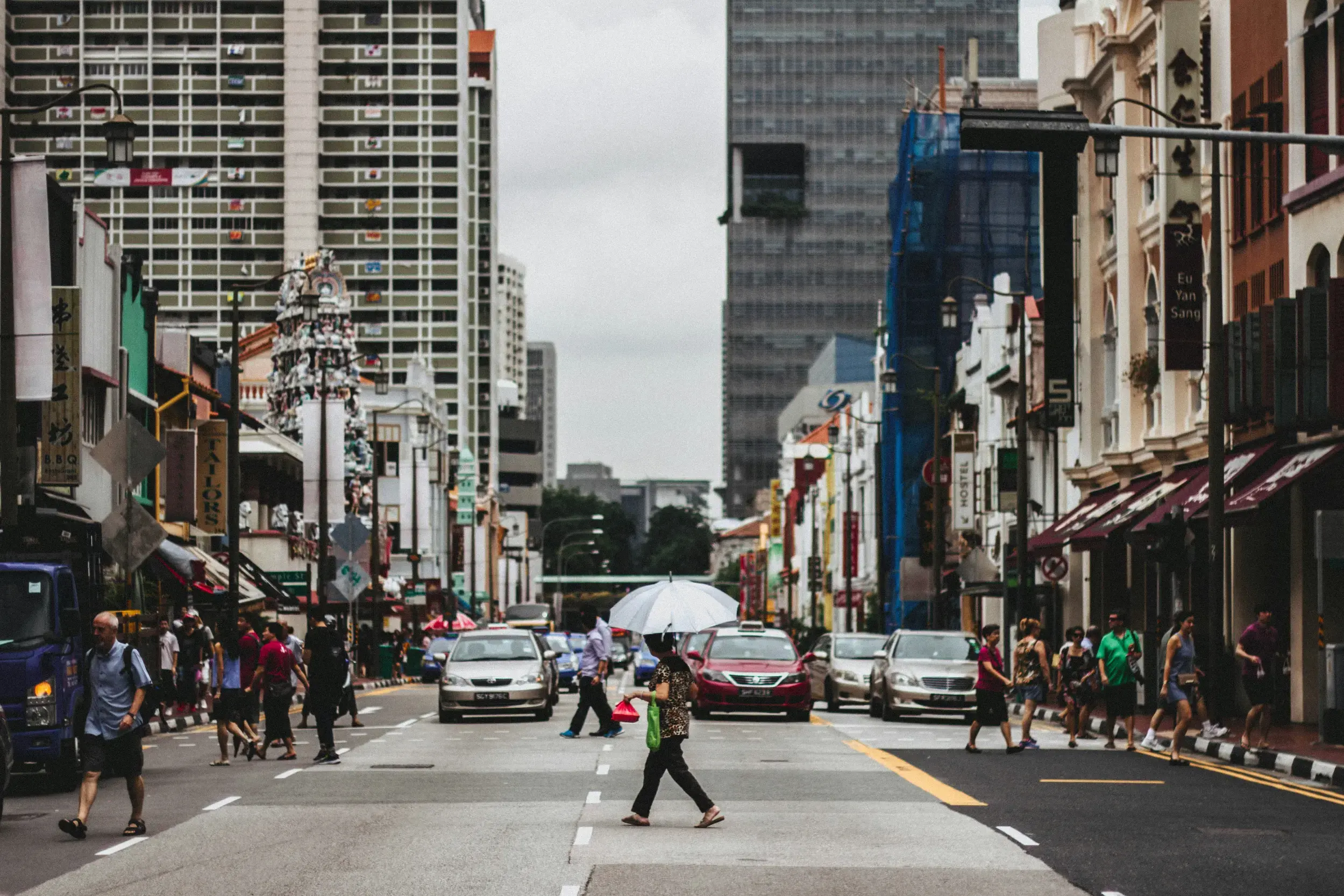 A person with an umbrella crosses a busy city street lined with diverse architecture, including traditional shophouses and a colorful Hindu temple. Pedestrians walk along the sidewalks past parked cars and vibrant shopfronts under an overcast sky.