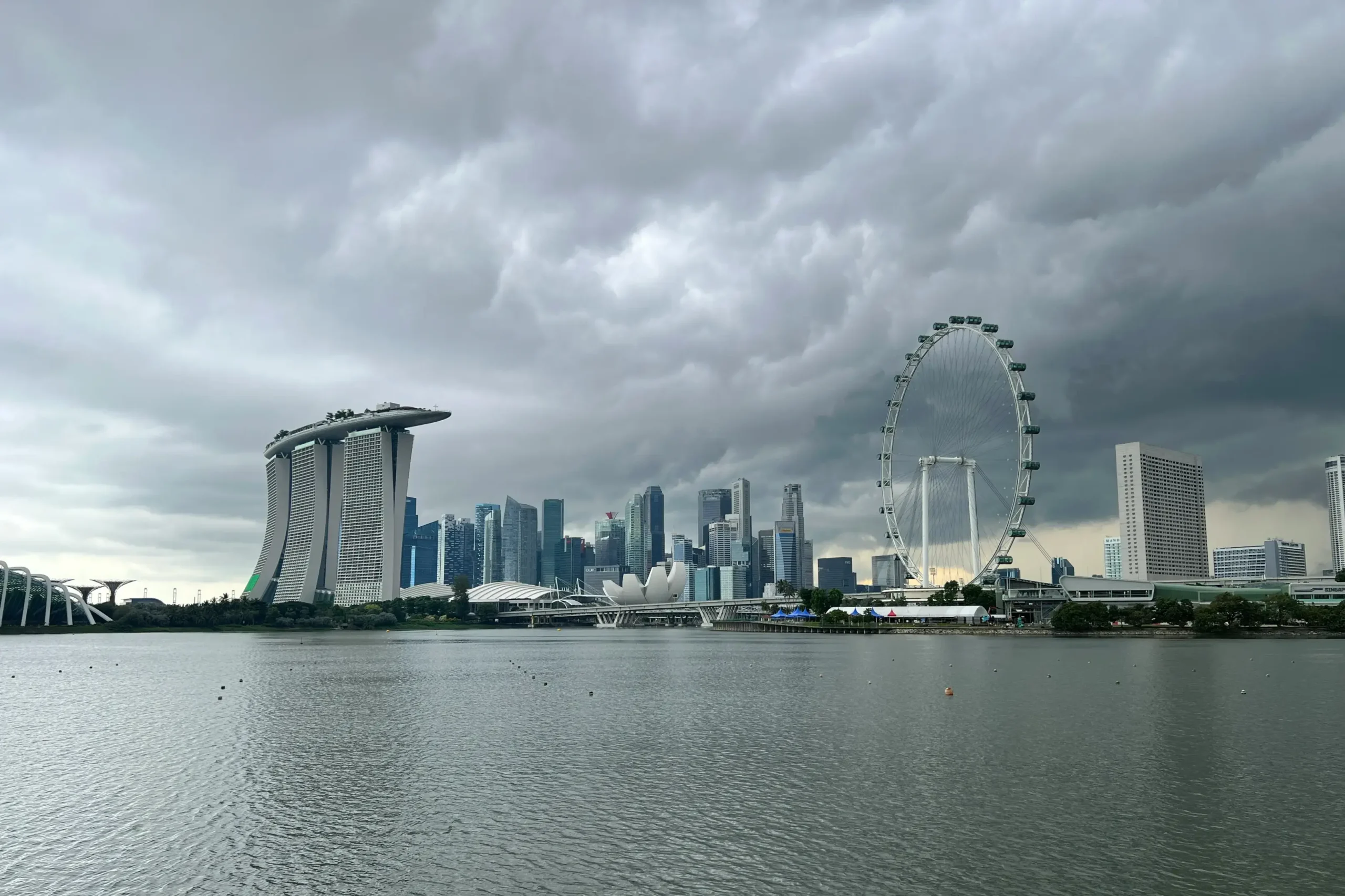 A wide shot captures the Singapore skyline under a heavy, overcast sky filled with dramatic grey storm clouds. The iconic Marina Bay Sands and Singapore Flyer stand prominently along the waterfront, reflecting the city's modern architectural style.