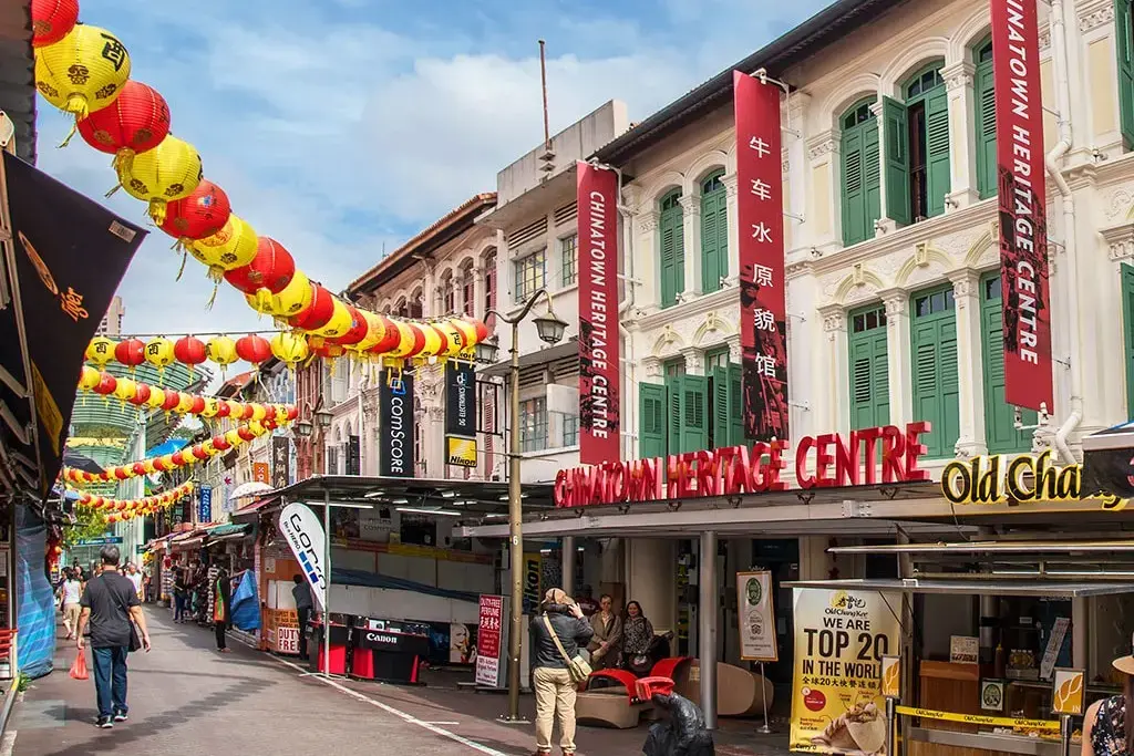 This image captures a vibrant street scene in Singapore's Chinatown, featuring the historic Chinatown Heritage Centre housed in beautifully restored shophouses with green shutters. Rows of red and yellow lanterns hang overhead, while pedestrians stroll past market stalls and local eateries like Old Chang Kee.