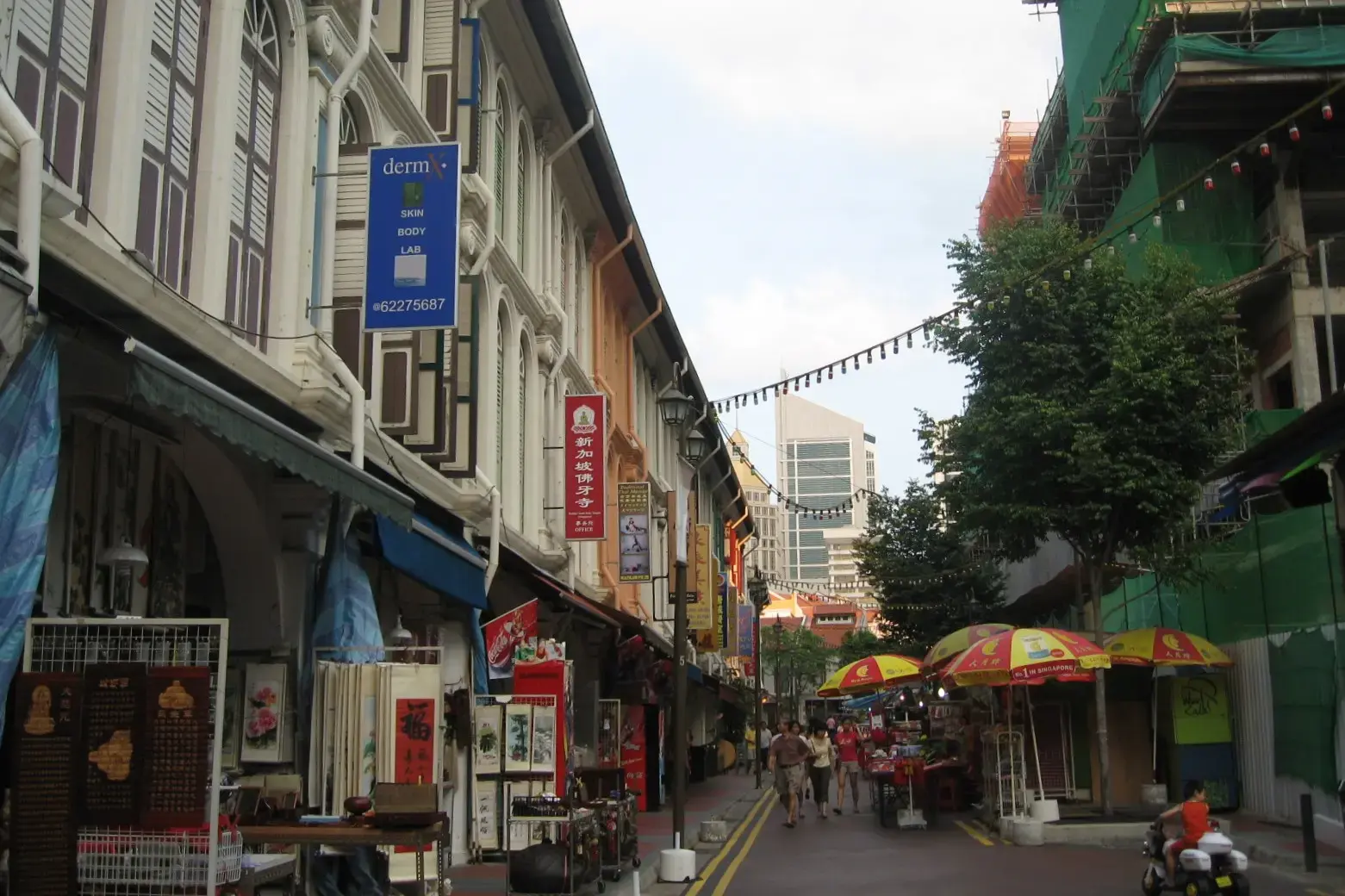 This street scene features a row of traditional shophouses with ornate facades and hanging signs alongside a bustling market area with colorful umbrellas. Pedestrians walk along the narrow road while modern skyscrapers and construction scaffolding rise in the background.