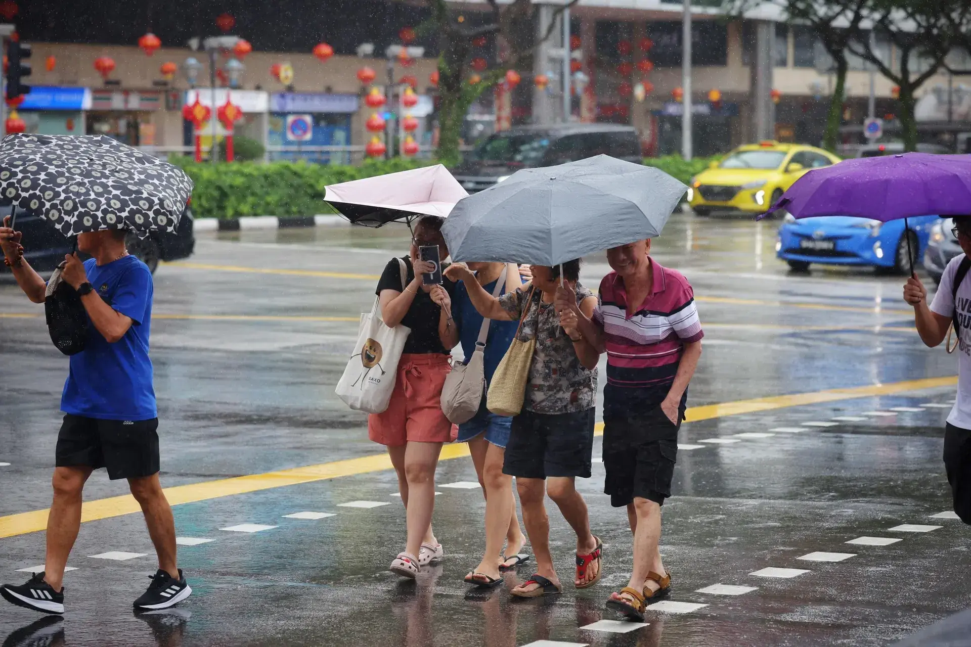 Pedestrians cross a rain-slicked city street while holding a variety of colorful umbrellas to shield themselves from the downpour. In the background, yellow taxis and red lanterns line the busy urban thoroughfare.