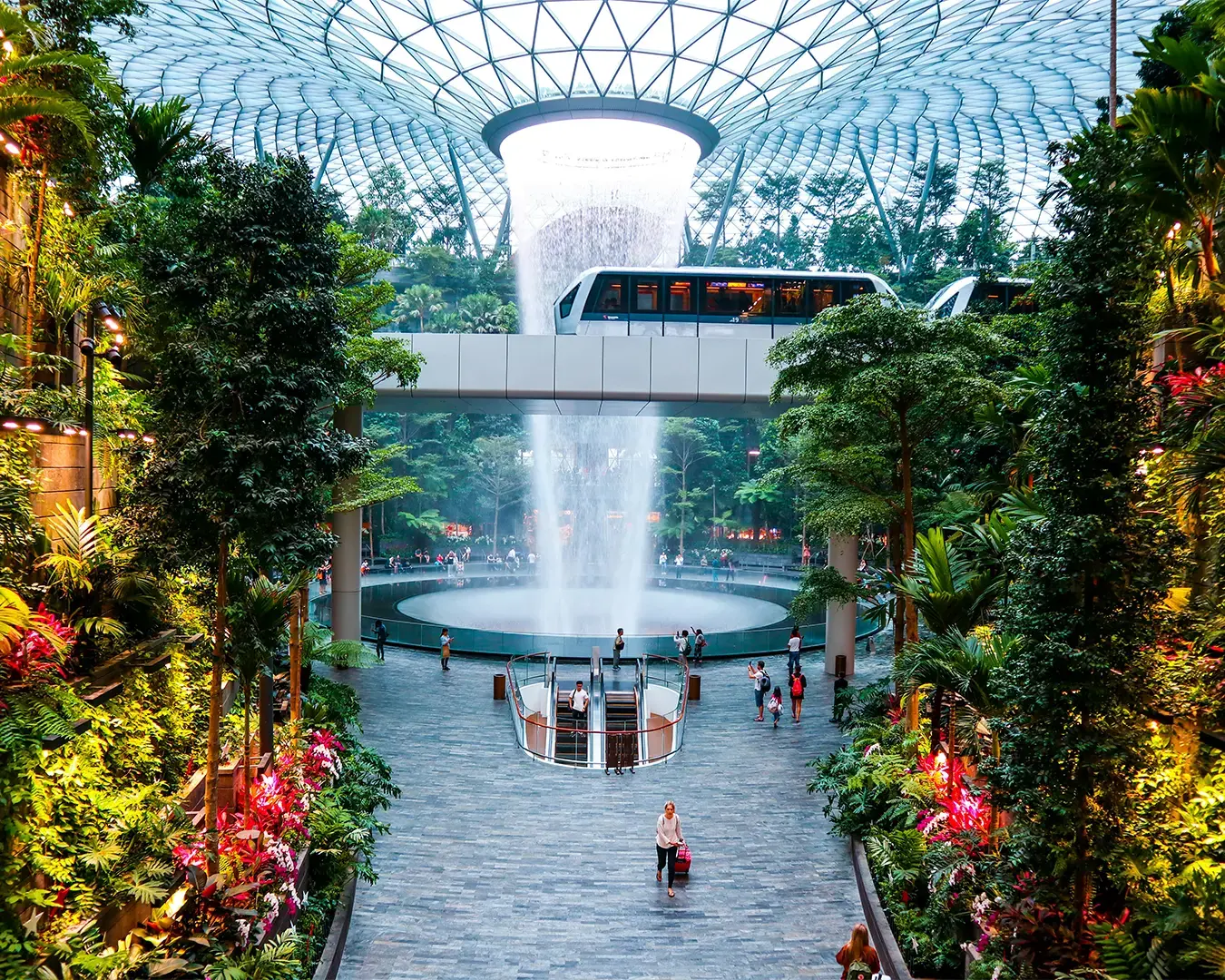 Indoor waterfall under a glass dome surrounded by lush greenery. A train crosses above, while people walk on a gray path, creating a serene, futuristic ambiance.