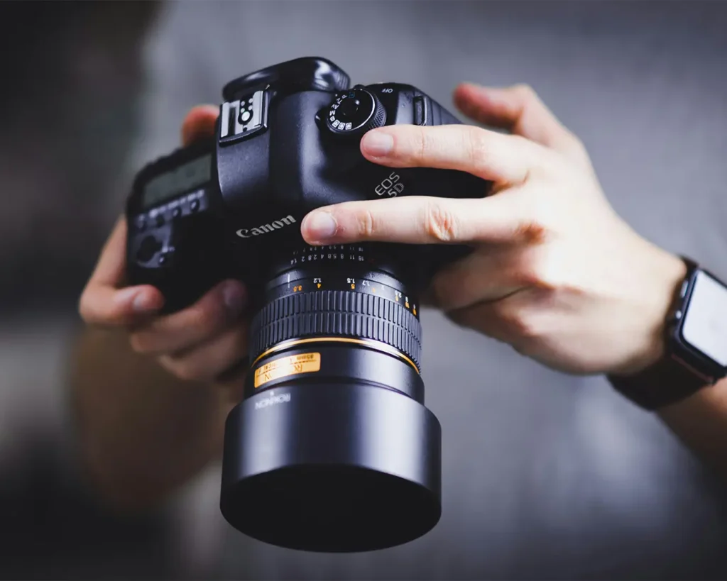 Close-up of hands holding a Canon EOS 5D DSLR camera with a large lens, showing precision and focus. The person wears a smartwatch. The tone is professional.