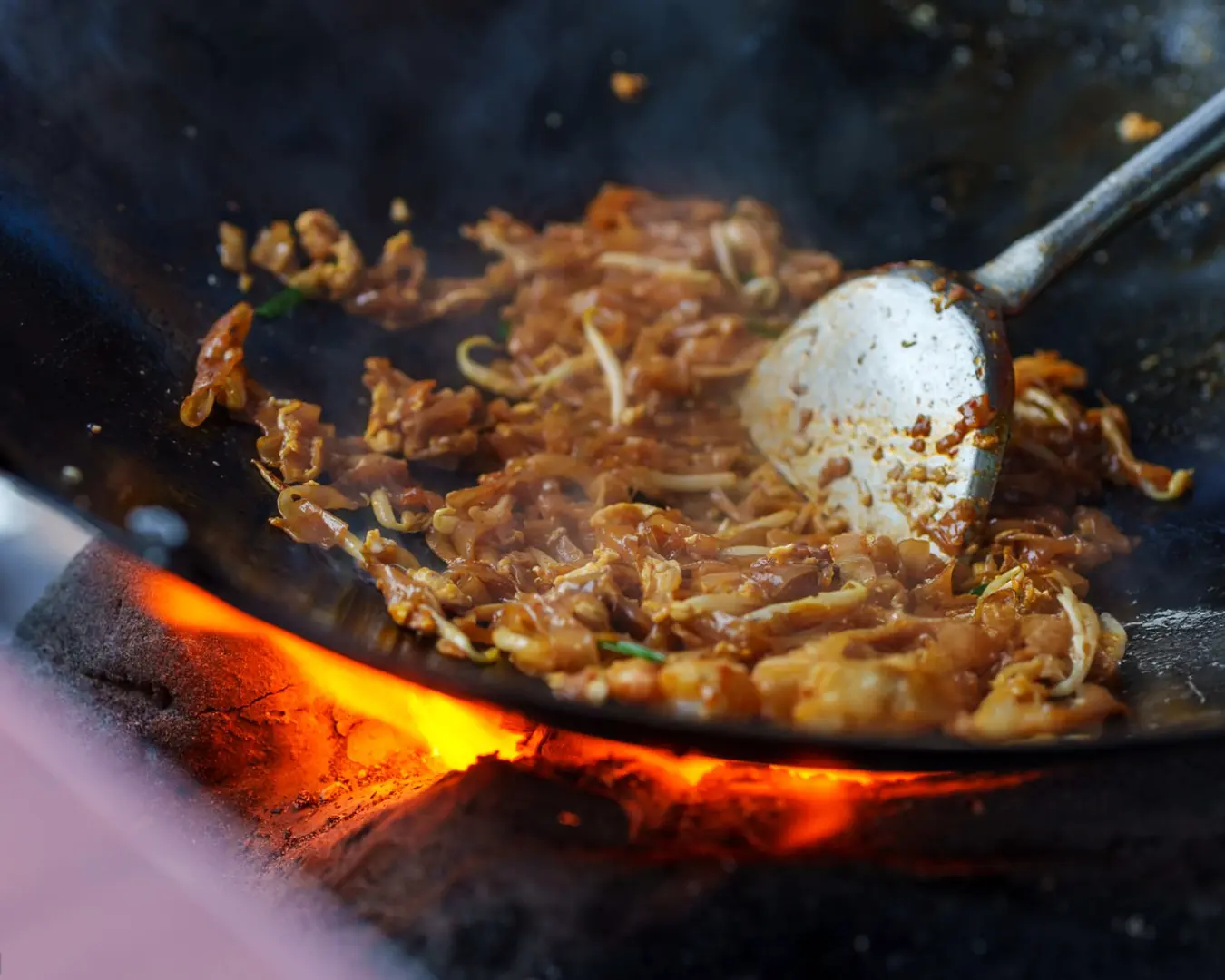 Stir-fried noodles with vegetables and chicken sizzling in a wok, vibrant flames beneath. A metal spatula stirs the dish, creating a dynamic, aromatic scene.