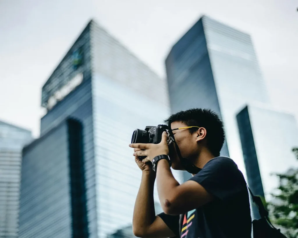 Young person taking a photo with a DSLR camera against a backdrop of tall, modern skyscrapers. The scene conveys a sense of focus and urban exploration.