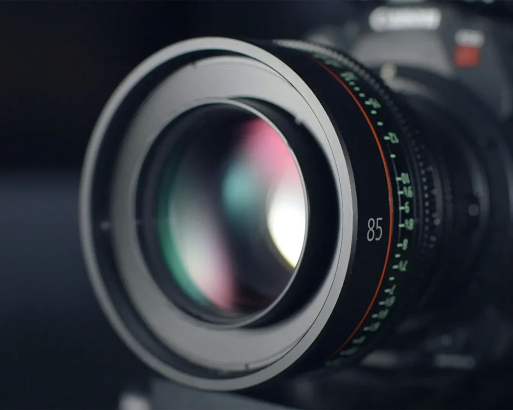 Close-up of a camera lens with a focal length of 85, featuring a sleek metallic frame. The lens reflects subtle colors, conveying precision and focus.