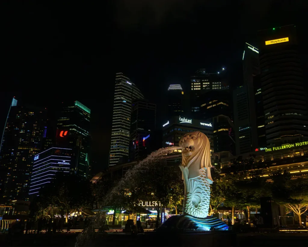 Night view of Singapore's Merlion statue, illuminated and spouting water, with a backdrop of towering, brightly lit city skyscrapers.