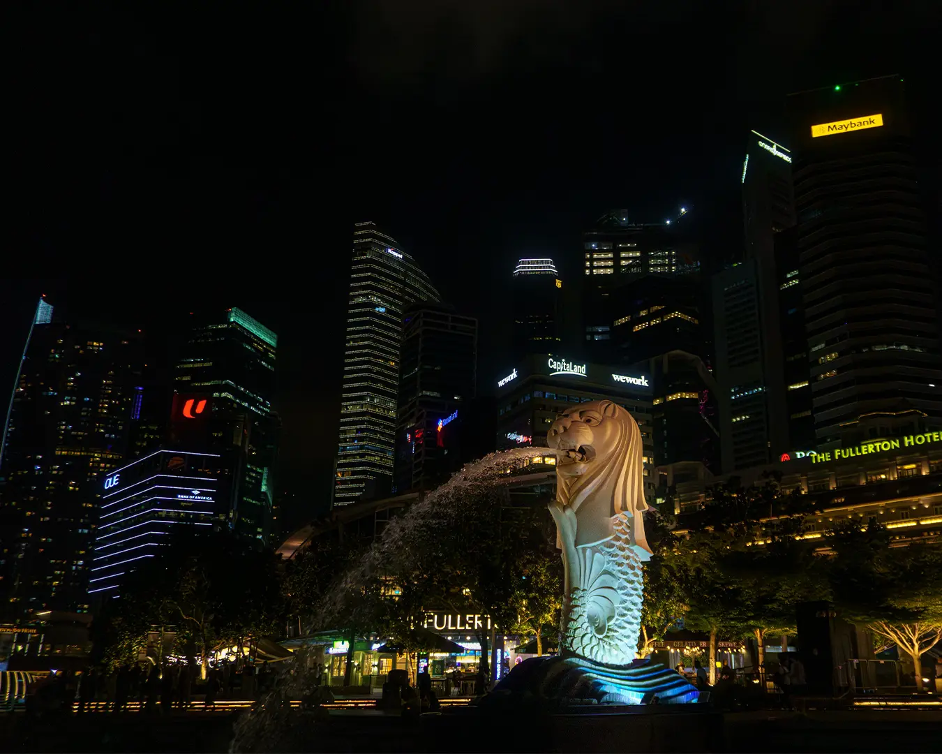 Night view of Singapore's Merlion statue, illuminated and spouting water, with a backdrop of towering, brightly lit city skyscrapers.