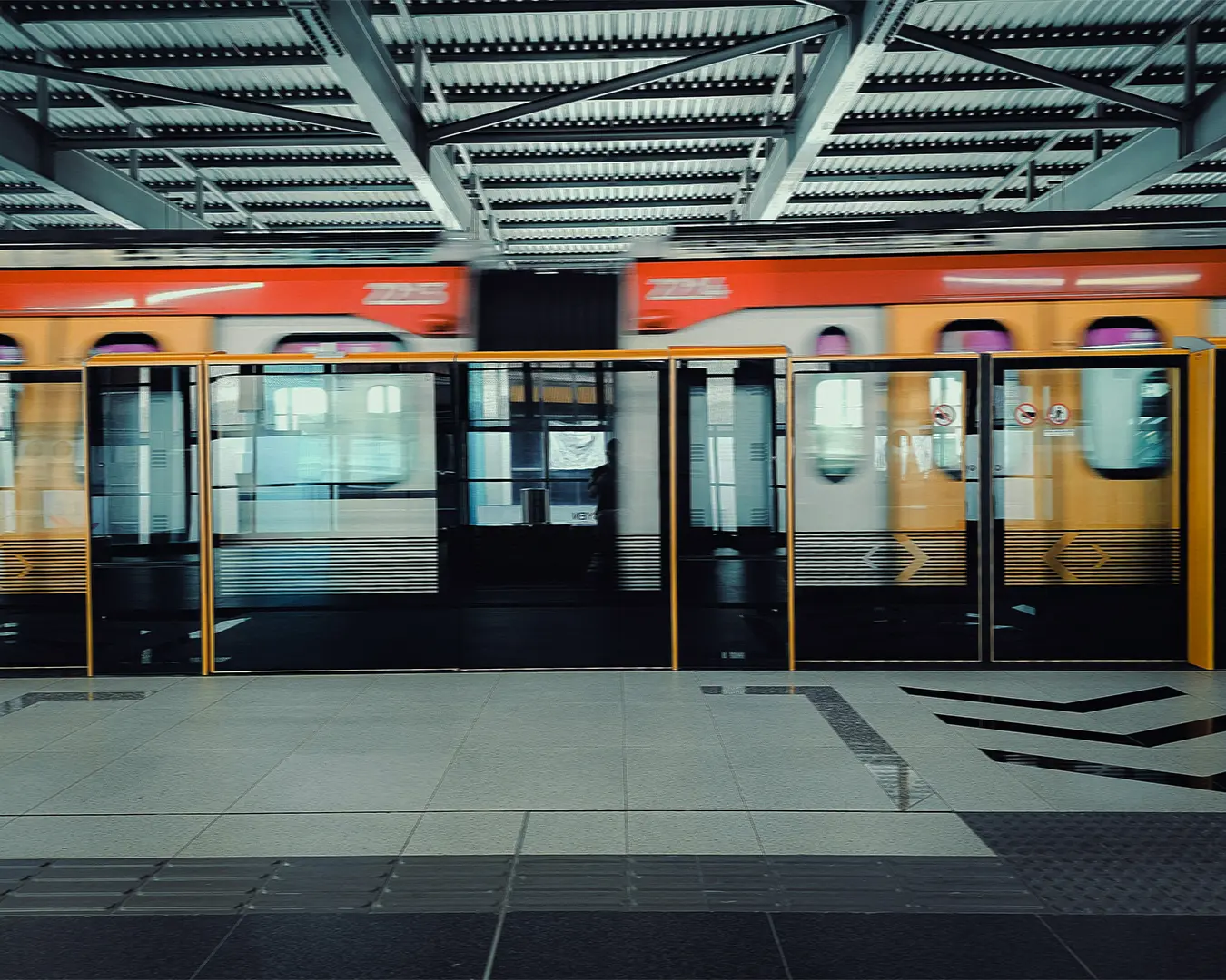 Blurry orange train moving past glass doors at a modern, empty station. The motion conveys speed and urban bustling atmosphere.