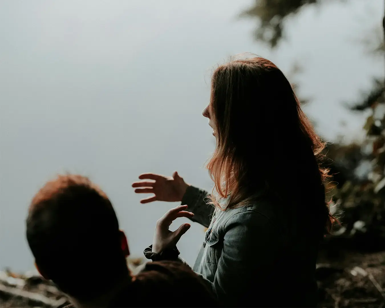 Two people in shadowed profile, one gesturing expressively with hands, near a serene, blurred water backdrop, conveying a contemplative mood.