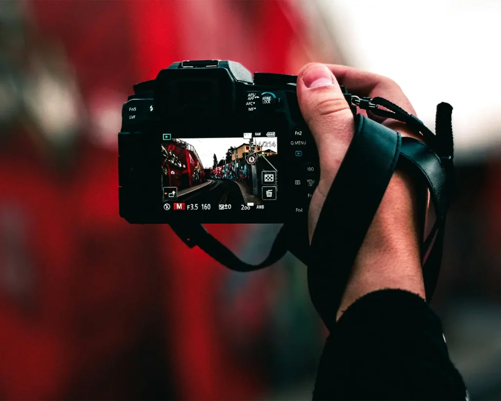 A hand holding a digital camera capturing a vibrant street scene with red walls. The camera's screen displays a clear image of the scene.
