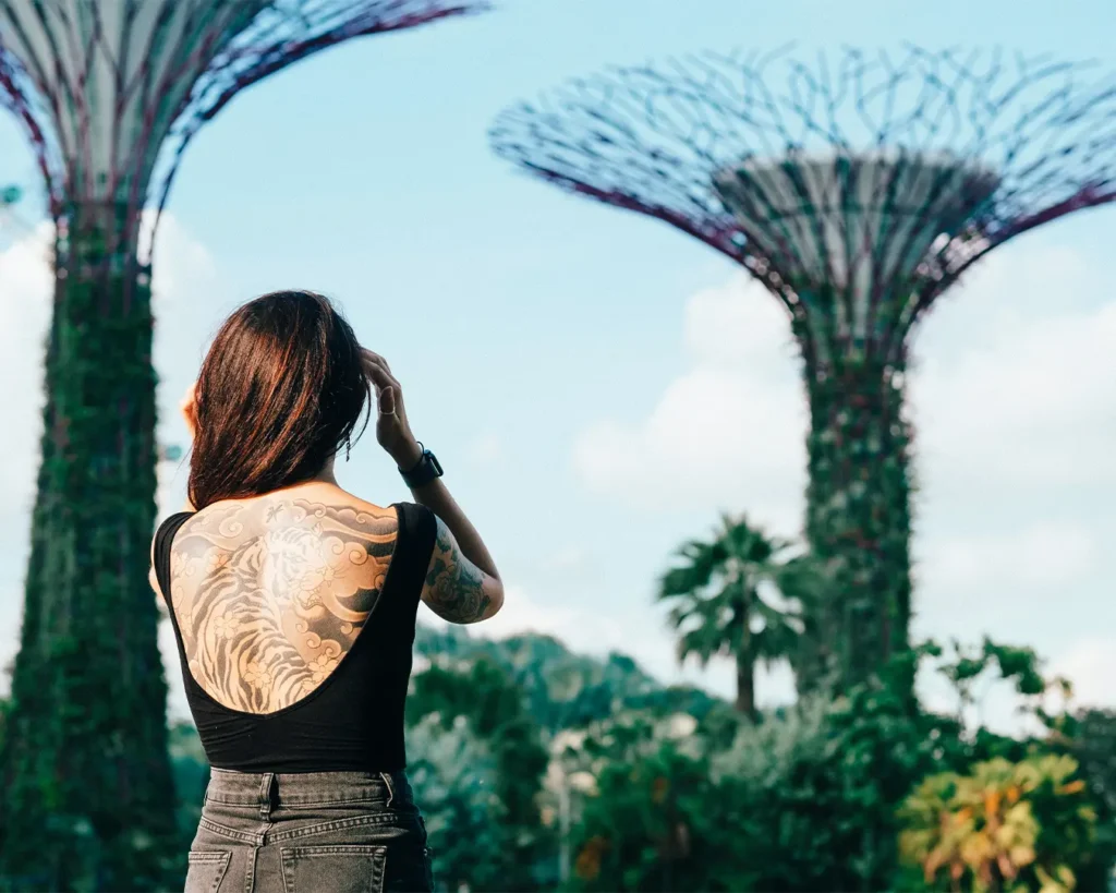 A woman with a large back tattoo wearing a black shirt stands outdoors, looking at towering futuristic structures against a bright blue sky.