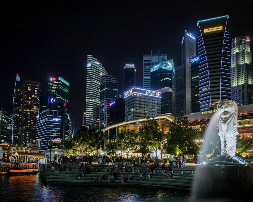 Night view of Singapore's skyline with illuminated skyscrapers. The iconic Merlion statue is in the foreground, surrounded by a lively crowd.