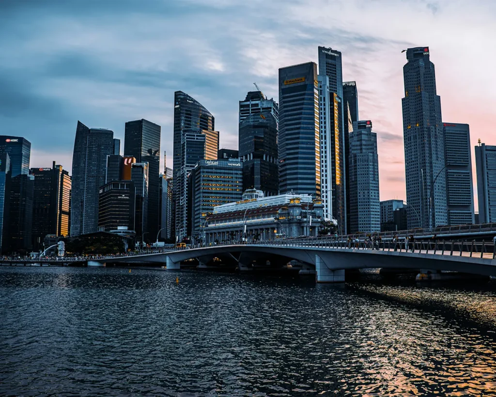 A skyline of tall skyscrapers under a cloudy blue sky at dusk, with a bridge crossing calm water in the foreground, creating a serene urban scene.