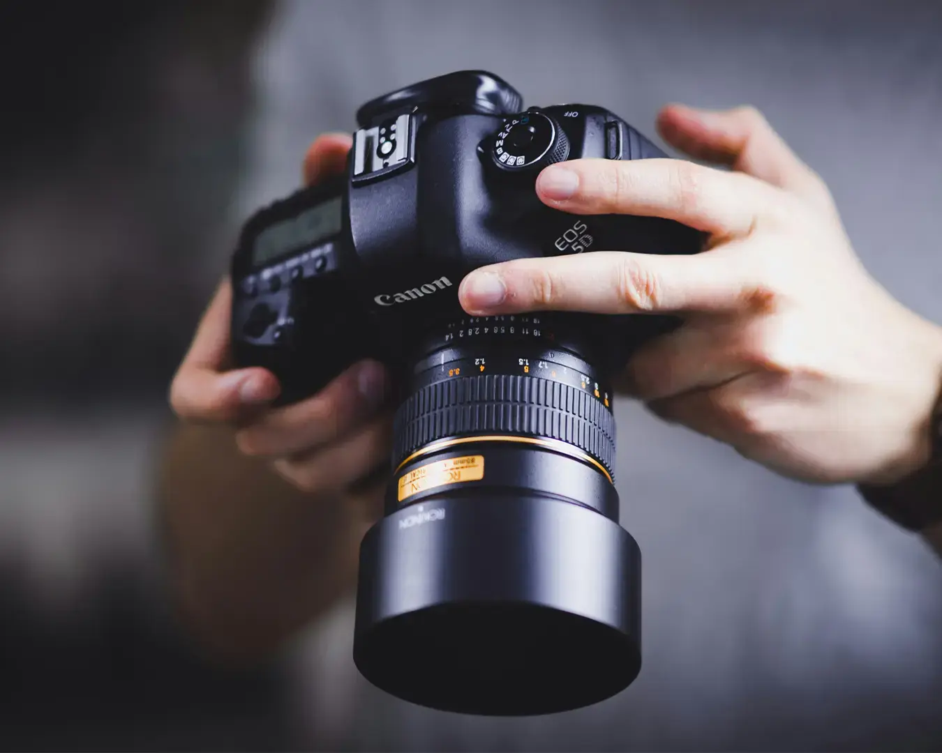A person holds a Canon EOS 5DS DSLR camera with a Samyang lens in both hands, focusing intently. The scene conveys concentration and precision.