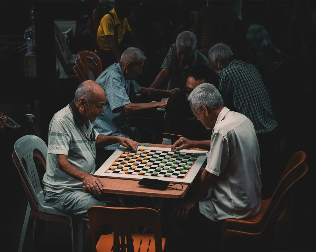 Elderly men focus intently on playing checkers at a wooden table in dim lighting. The atmosphere is contemplative, with others engaged nearby.