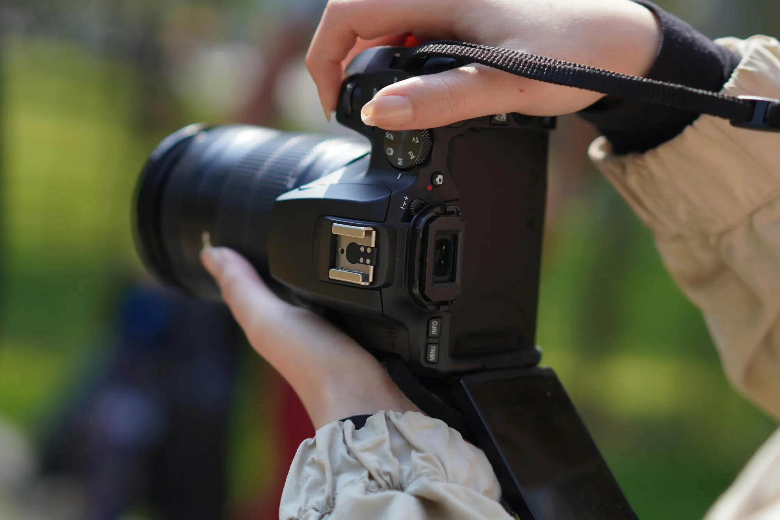 A person is holding a black DSLR or mirrorless camera with both hands, positioned to take a photo. The camera features a large lens and a flip-out screen, set against a softly blurred green outdoor background.