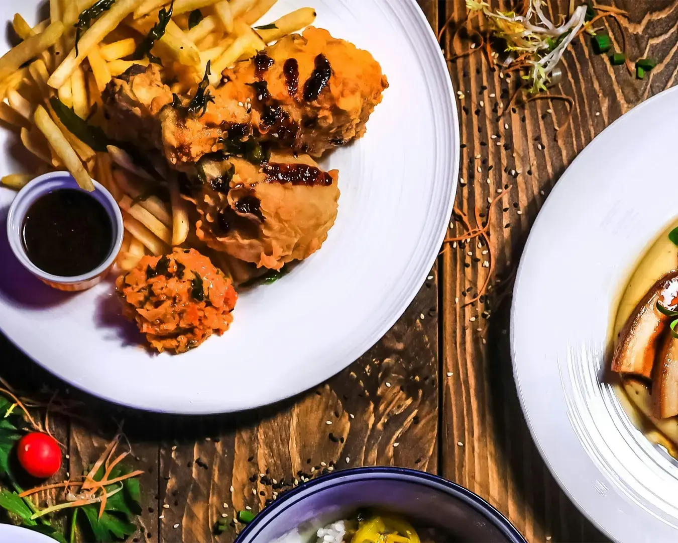 Plate with fried chicken, fries, and a dipping sauce on a wooden table. Nearby are garnishes, a bowl, and part of another dish, creating a vibrant, appetizing scene.