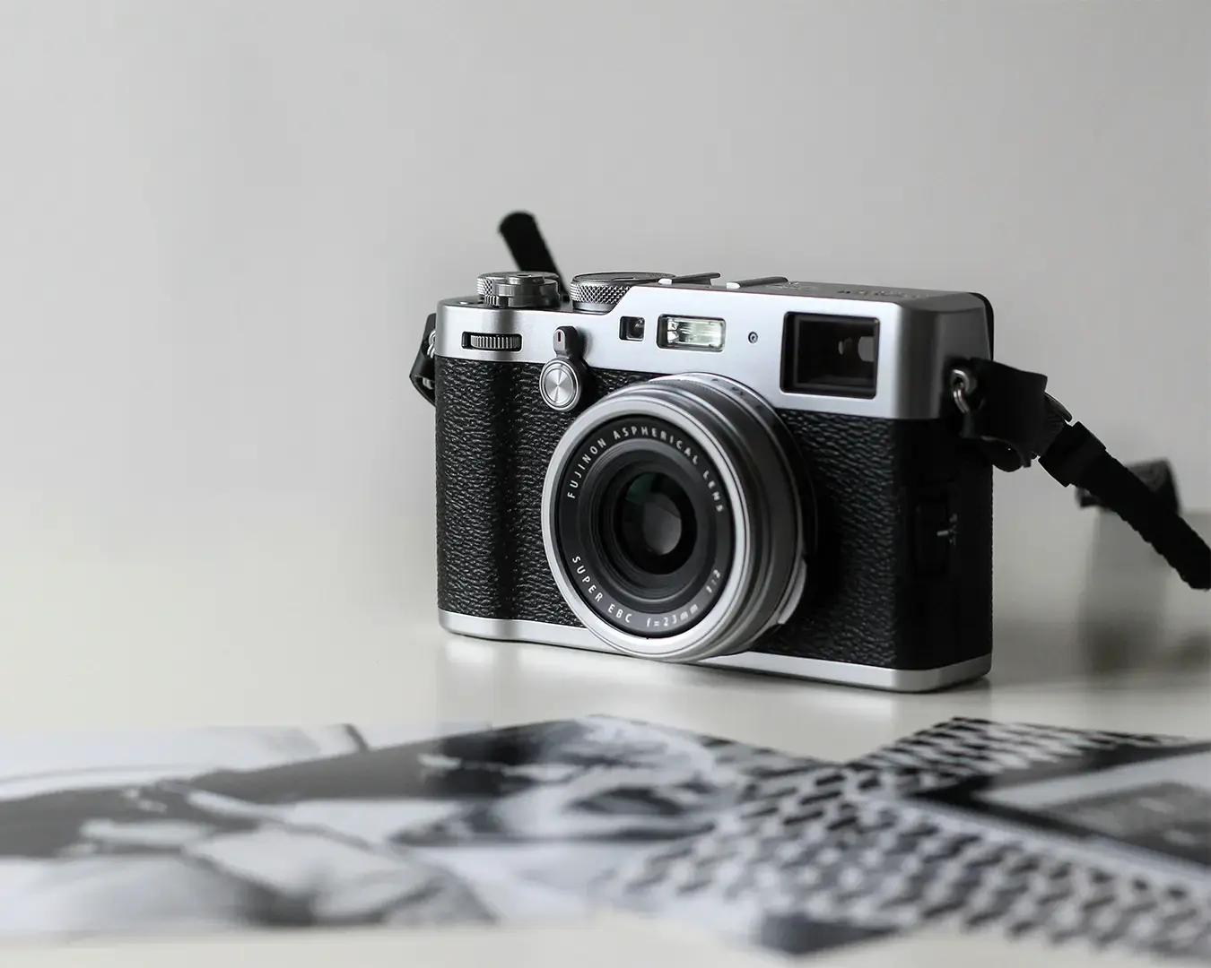 A vintage-style camera with a black textured body and silver lens sits on a white surface. In the foreground, blurred black-and-white photos are scattered.