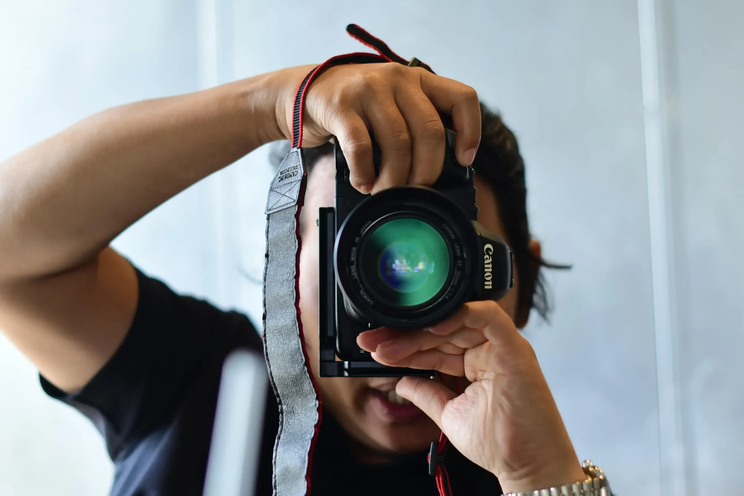 A person holds a Canon DSLR camera directly in front of their face, capturing a first-person perspective of someone taking a photograph. The camera lens features a vibrant green reflection, and a red and grey neck strap hangs down from the camera body.