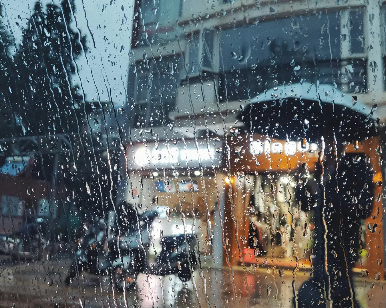 Rain-soaked window with water droplets blurring a city street view. A person with an umbrella walks past a lit-up store, evoking a moody, calm ambiance.