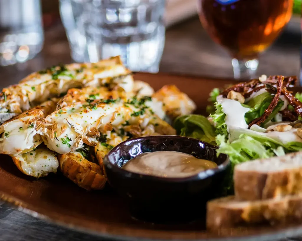 Grilled lobster tails garnished with herbs on a wooden plate, accompanied by fresh salad, dipping sauce in a black bowl, and slices of bread.