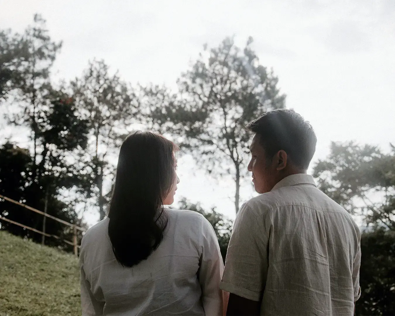 Two people stand outdoors, backs facing the camera, looking at each other. They're surrounded by trees under a bright sky, creating a serene, intimate mood.