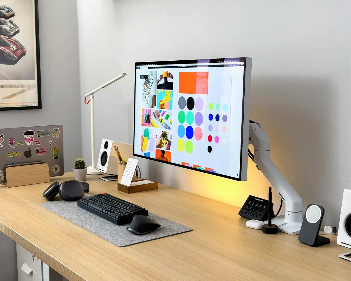 Modern desk setup featuring large monitor displaying colorful designs, a keyboard, mouse, and a laptop with stickers. Bright, organized workspace with a sleek, minimalist vibe.