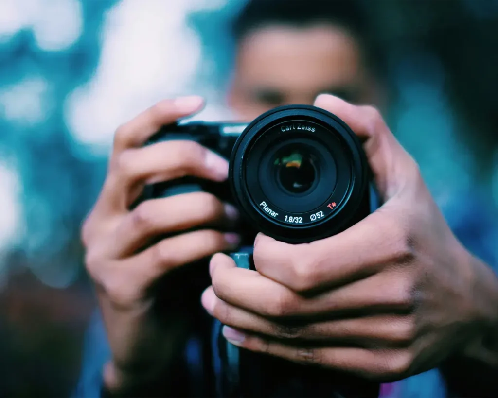 Close-up of a person holding a camera, focusing on the lens with fingers wrapped around it. The background is softly blurred, creating a dynamic feel.