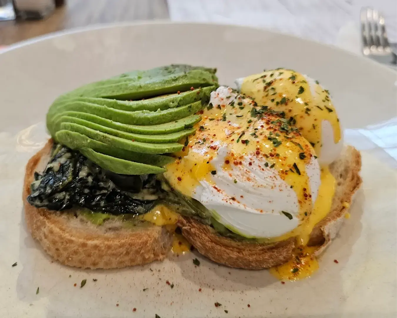 Open-faced sandwich with sliced avocado, poached eggs, and Hollandaise sauce on toasted bread. Topped with spices, it appears fresh and appetizing.