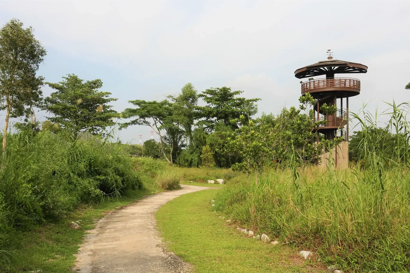 A winding dirt path leads through lush green grass and trees toward a unique, multi-level observation tower with a spiral staircase. The architectural structure stands tall against a pale sky, overlooking a peaceful, natural landscape.