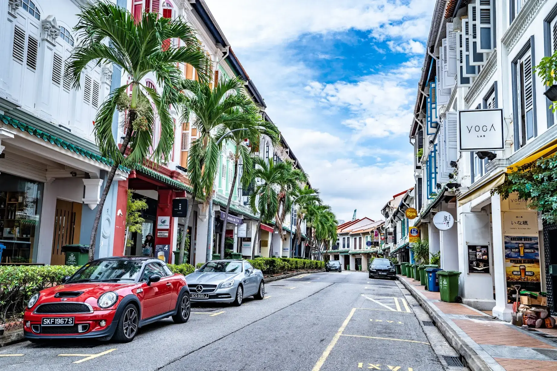 A vibrant street is lined with colorful, historic shophouses and tall palm trees under a bright, cloudy sky. Several cars are parked along the road, which features businesses like a yoga studio and local eateries nestled within the traditional architecture.
