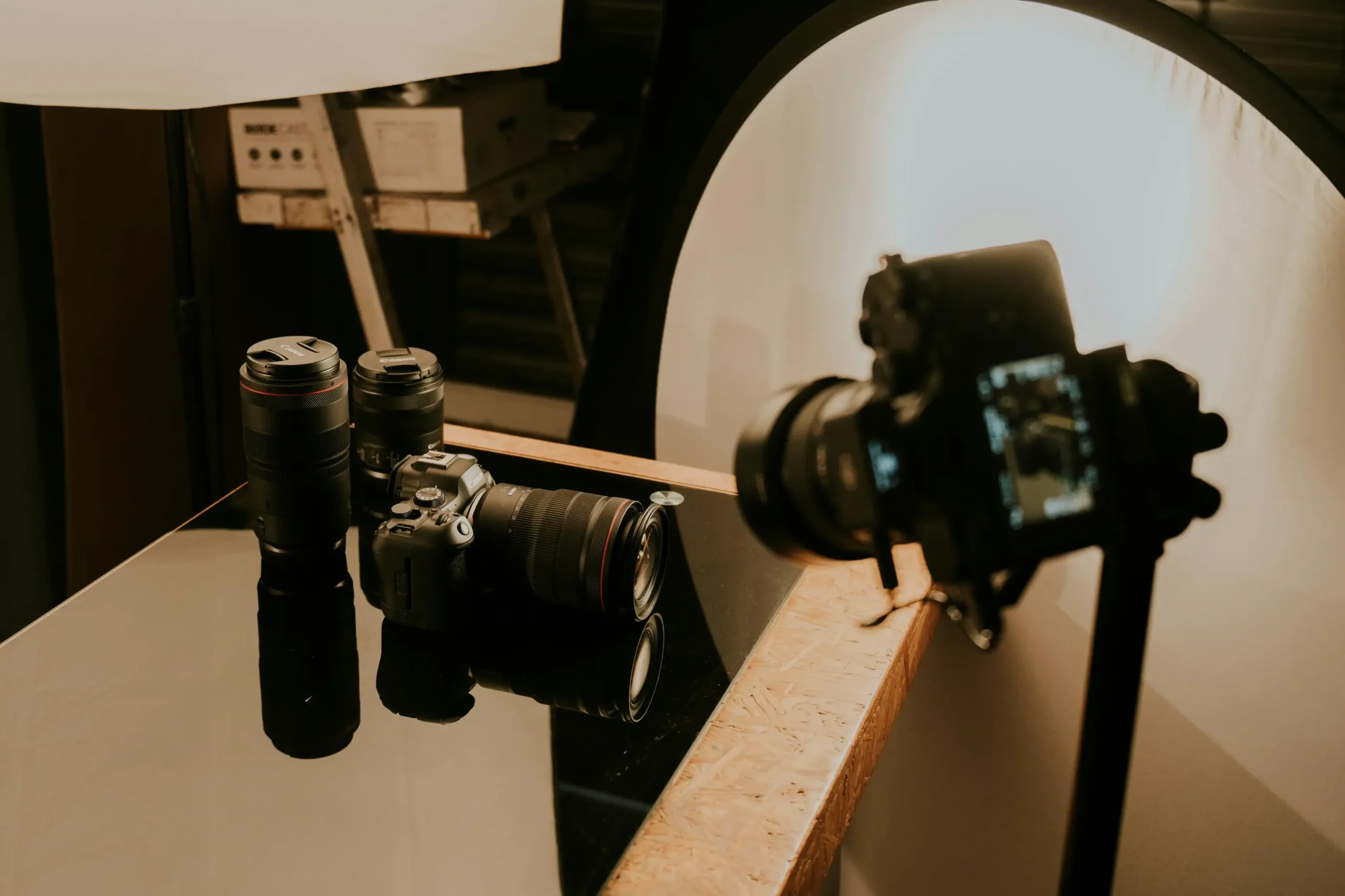 A professional camera setup sits on a reflective black surface, featuring multiple lenses and a mirrorless body positioned for a product shoot. Large softbox lighting and a circular reflector illuminate the gear, highlighting the sleek design of the photography equipment.